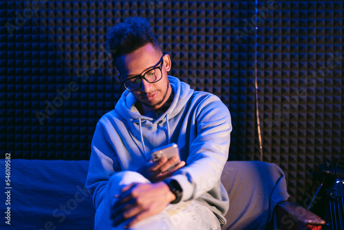 Close-up portrait of an attractive curly guy sitting on sofa using smartphone browsing web service at modern loft industrial style interior splashed by blue light
