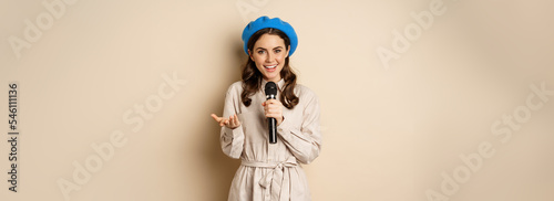 Portrait of happy stylish woman performing, singing with microphone, posing against beige background