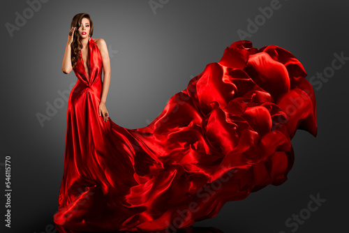 Sexy Fashion Model in Red Silk Dress. Glamour Woman in Long Luxury Gown flying on Wind with Wavy Hairstyle over Dark Gray Background. Elegant Lady dancing