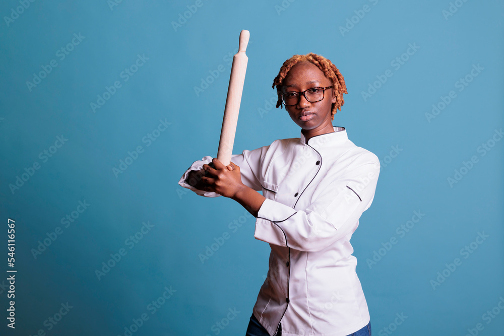African american female baker holding rolling pin simulating going to bat ball while looking at camera in studio shot with blue background. Serious female kitchen staff wearing work uniform. African american female baker holding rolling pin simulating going to bat ball while looking at camera in studio shot with blue background. Serious female kitchen staff wearing work uniform.