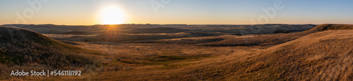 Wide panorama of a sunset over a dry badlands setting with the sunlight highlighting hills under a clear sky. 