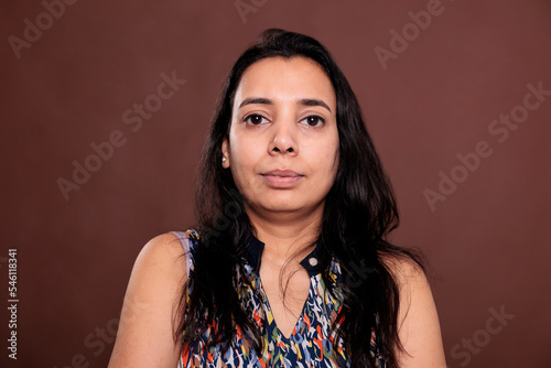Indian woman looking at camera medium closeup portrait. Calm lady with neutral facial expression close view, relaxed smiling person front view studio medium shot on brown background
