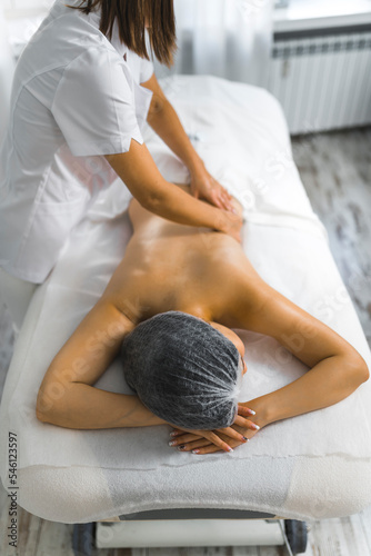 Vertical indoor shot of skinny young Asian woman lying on her stomach on massage bed being massaged by unrecognizable caucasian female massage therapist. SPA day. High quality photo