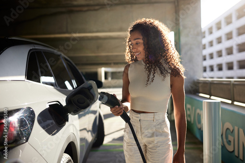 Young woman holding electric plug by car at charging station