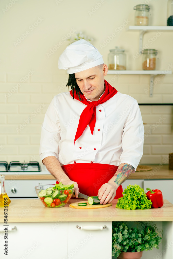 Young handsome chef cutting green cucumber on wooden board in kitchen. Positive guy with dreadlocks in uniform preparing vegetable salad. Young handsome chef cutting green cucumber on wooden board in kitchen. Positive guy with dreadlocks in uniform preparing vegetable salad.