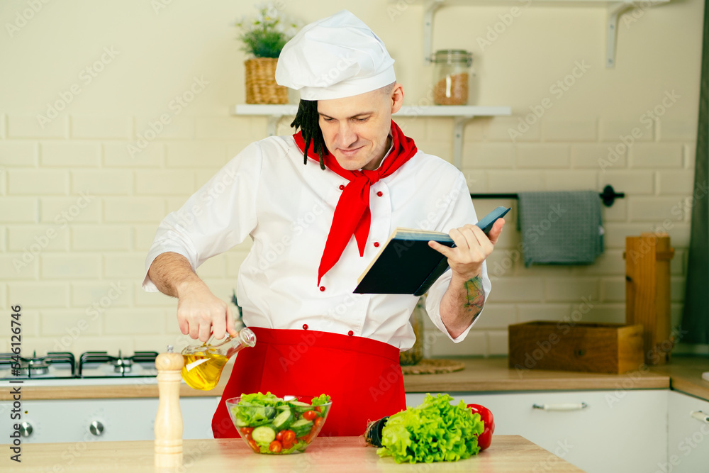 Young man dressed as chef pouring olive oil on vegetable salad, reading recipe book in kitchen. Concept of food, cooking, healthy lifestyle. Young man dressed as chef pouring olive oil on vegetable salad, reading recipe book in kitchen. Concept of food, cooking, healthy lifestyle.