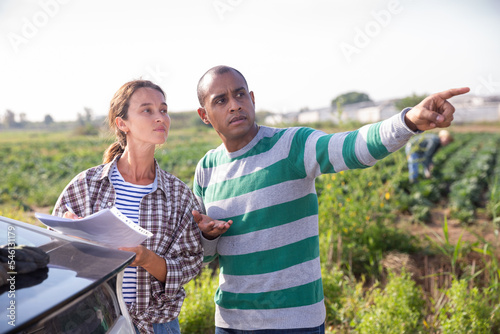 Female farmer discussing some papers with hispanic partner while standing near car on farm field © JackF