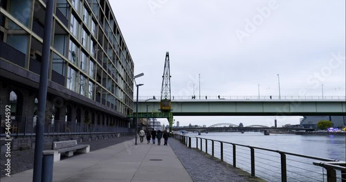 Reveal wide tilt up shot of a pedestrian walkway along the rhine river in cologne germany with pedestrians, severins bridge and modern building on the side with view of the hohenzollern bridge photo