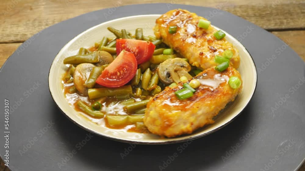 Cutlets with green beans and mushrooms in tomato sauce garnished with tomatoes and green onions in a plate rotate in a circle. Medium plan Cutlets with green beans and mushrooms in tomato sauce garnished with tomatoes and green onions in a plate rotate in a circle. Medium plan
