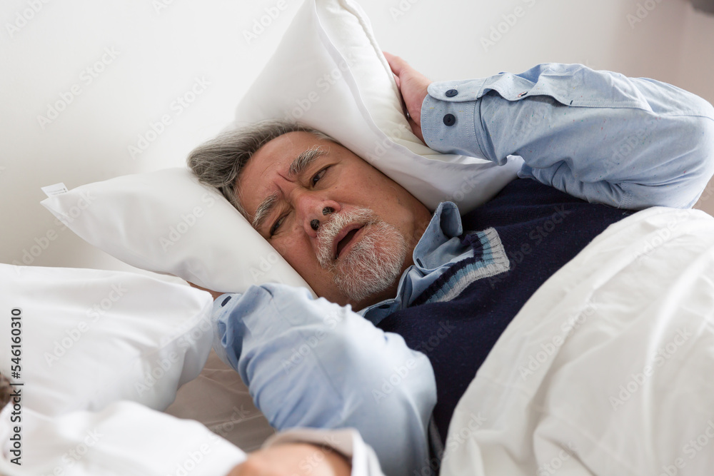 Elderly Asian man having trouble sleeping squeezes white pillow around ears for peace and quiet on bed in bedroom - obrazy, fototapety, plakaty 
