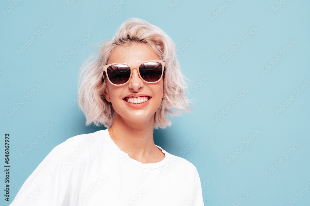 Young beautiful smiling female in trendy summer clothes. Sexy carefree woman posing near blue wall in studio. Positive blond model having fun and going crazy. Cheerful and happy. In sunglasses Young beautiful smiling female in trendy summer clothes. Sexy carefree woman posing near blue wall in studio. Positive blond model having fun and going crazy. Cheerful and happy. In sunglasses