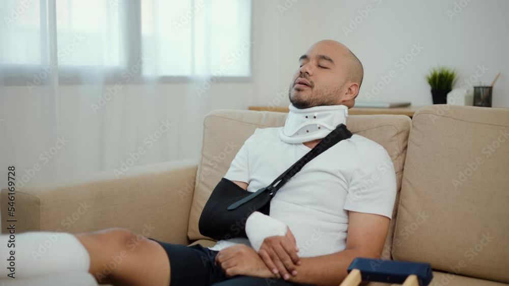 Happy man recovery from accident fracture broken bone injury with leg splints in cast, neck splints collar, arm splints, sling support arm in living room. Social security and health insurance concept.