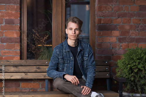 Portrait of a young Caucasian guy with a stylish hairstyle in a denim jacket who is sitting on a bench.