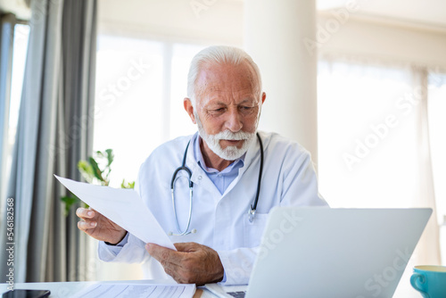 Serious old mature professional male doctor using laptop computer in hospital office having medical webinar training, writing in healthcare report, consulting patient online at telemedicine meeting.