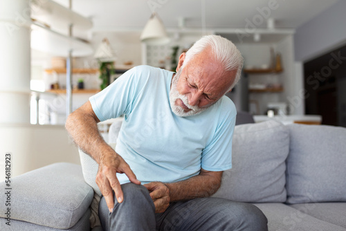 The older man is sitting on the couch at home, has pain in the knee joint, holding his leg, osteoarthritis concept.