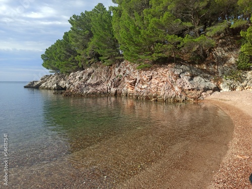 Beautiful landscape of a Kamenovo pebble beach located in a bay surrounded by green mountains near village of Rafailovici at blue sky background on a sunny summer morning. Budva Riviera. Montenegro.