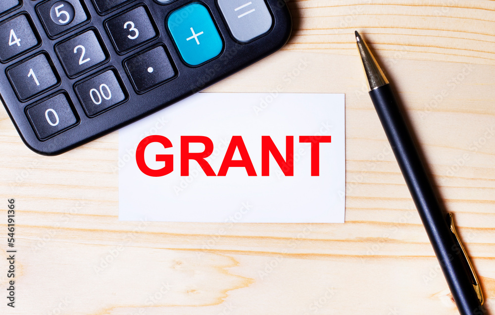 On a light wooden background, a black calculator, a pen and a business card with the text GRANT On a light wooden background, a black calculator, a pen and a business card with the text GRANT