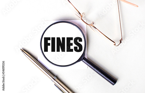 Golden pen and glasses, black magnifying glass with the text FINES on a light background.