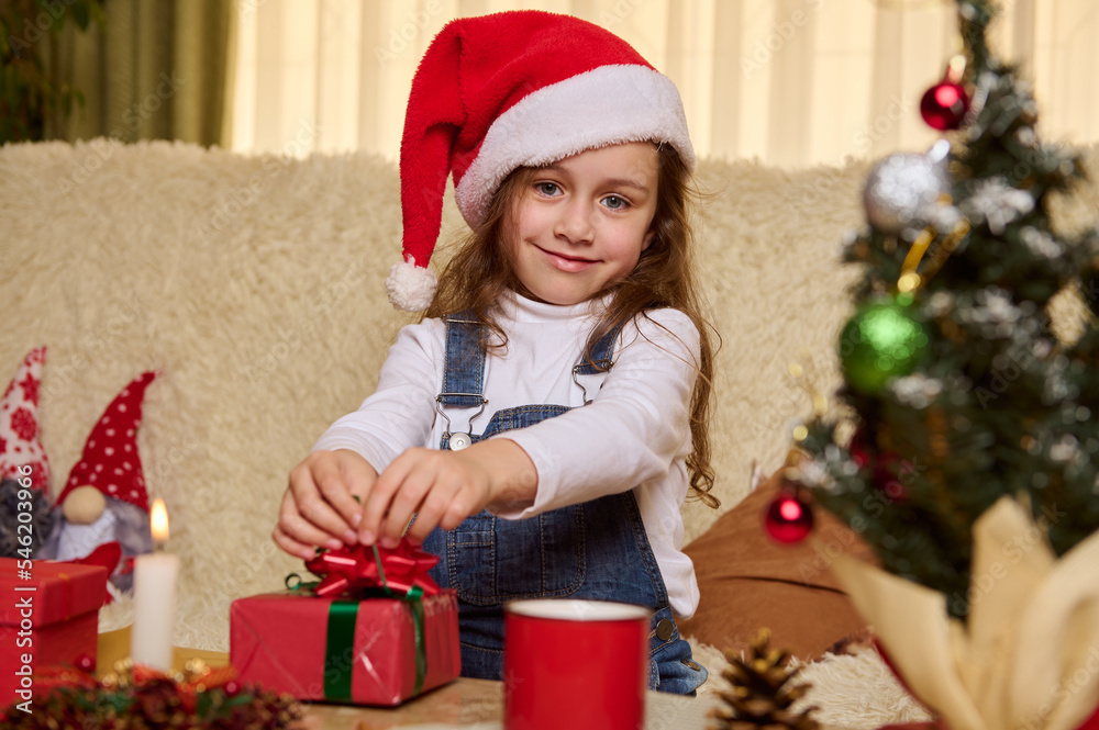 Delightful child, adorable 5 years old little girl cutely smiles looking at camera, sitting on sofa, unpacking Christmas gift in cozy home interior with blurred Christmas tree on foreground. New Year Delightful child, adorable 5 years old little girl cutely smiles looking at camera, sitting on sofa, unpacking Christmas gift in cozy home interior with blurred Christmas tree on foreground. New Year