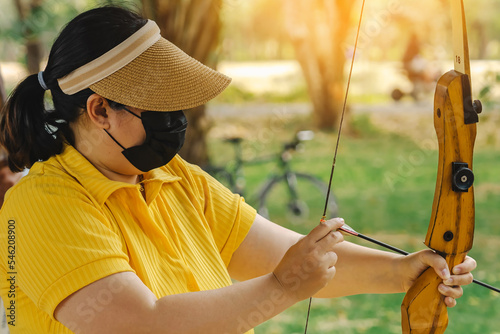 Asian woman wear visor with face mask aims archery bow and arrow to colorful target in shooting range during training and competition. Exercise and concentration with outdoor archery.Sport,Recreation