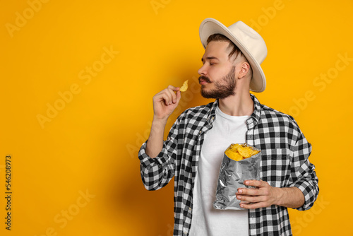Canvas-taulu Handsome young man with bag of tasty potato chips on yellow background