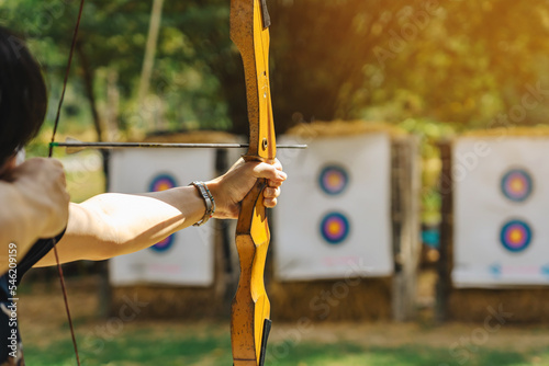 Hands of woman aims archery bow and arrow to colorful target in shooting range during training. Exercise and concentration with outdoor archery. Selective focus on hand. Sport, Recreation concept. photo