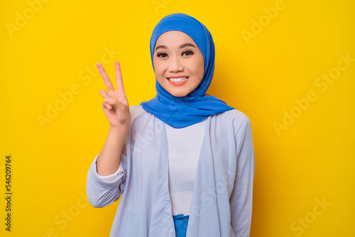 Portrait of cheerful young Asian Muslim woman showing peace gesture isolated over yellow background © Sewupari Studio