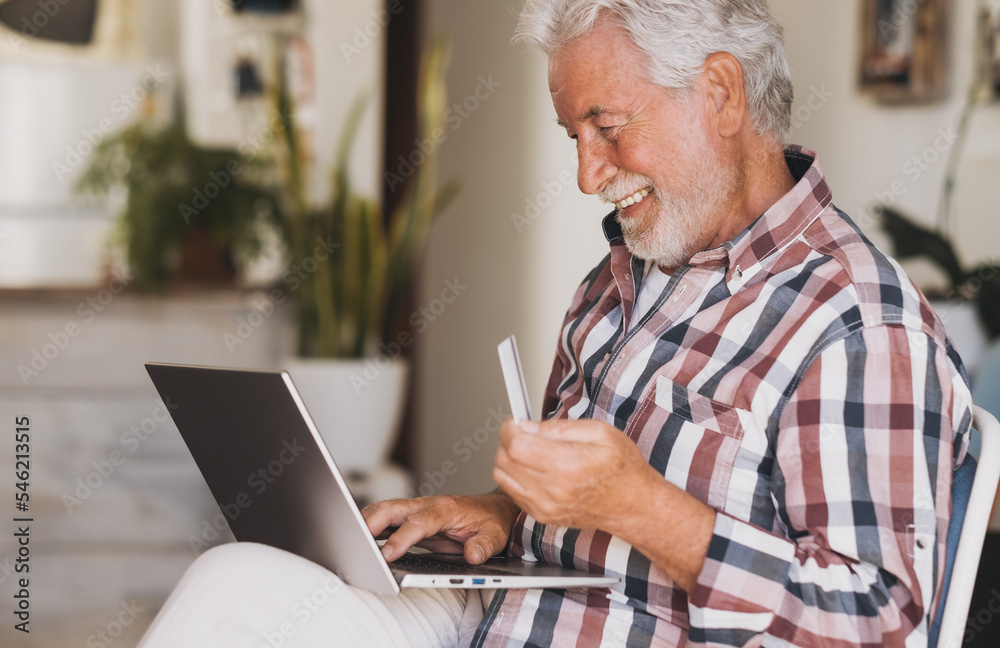 Elderly bearded man browsing by laptop enjoying black friday shopping online. Joyful and smiling senior male at home being in great mood using credit card for e-commerce purchases Elderly bearded man browsing by laptop enjoying black friday shopping online. Joyful and smiling senior male at home being in great mood using credit card for e-commerce purchases