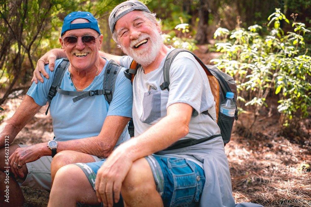 Couple of happy senior men in mountain excursion embracing looking at camera laughing while sitting to rest, enjoying healthy lifestyle in nature Couple of happy senior men in mountain excursion embracing looking at camera laughing while sitting to rest, enjoying healthy lifestyle in nature