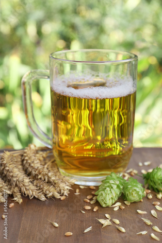 Mug with beer, fresh hops and ears of wheat on wooden board outdoors