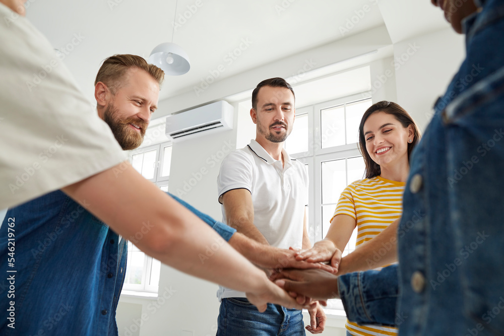 Fototapeta premium Team of cheerful people joining hands to show concept of teamwork and achieving success together. Group of happy men and women in casual T shirts standing in office, putting hands together and smiling