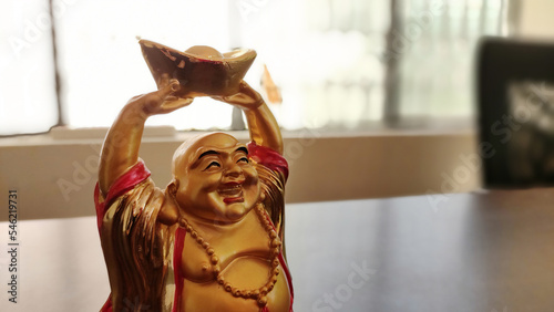 A golden Laughing Buddha statue holding a gold ingot with his hands upright. The statue is smiling broadly and wearing a red robe with a beaded necklace. photo