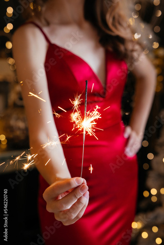 a girl in a red silk tight-fitting dress stands in a dark room against the background of New Year's decorations and holds a sparkler in her hands