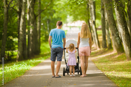 Young adult parents and little daughter pushing baby stroller and walking at tree alley of park in warm sunny summer day. Spending time together and breathing fresh air. Two child family. Back view. photo