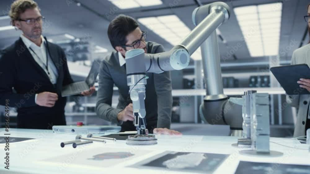 Industrial Robotics Engineer Talking and Discussing the Development of a Robotic Arm Machine with His Colleagues. Scientists with Laptop and Tablet Computers Research the Possibilities of the Robot. Industrial Robotics Engineer Talking and Discussing the Development of a Robotic Arm Machine with His Colleagues. Scientists with Laptop and Tablet Computers Research the Possibilities of the Robot.