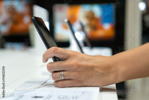 Close-up view shows the hand of a female customer choosing a black smartphone in a mobile phone store. Difficult decision, different choice