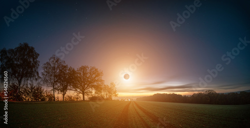 total solar eclipse during sunrise over a green field and trees along the road