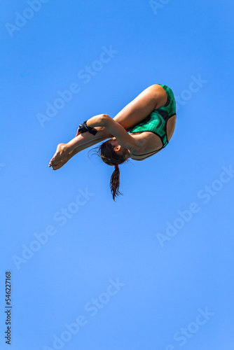 Diving Girl Athlete Unrecognizable Somersault High Action Off High Platform Against Blue Sky Into Swimming Pool.