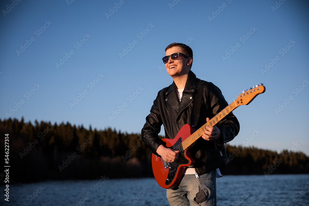 Naklejka premium a guy with an electric guitar smiling plays against the background of nature, a musician guitarist in sunglasses and a leather jacket