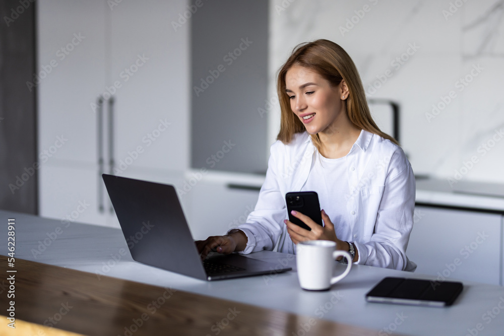 Pretty young woman having breakfast while sitting at the kitchen table, working on laptop computer, using mobile phone Pretty young woman having breakfast while sitting at the kitchen table, working on laptop computer, using mobile phone