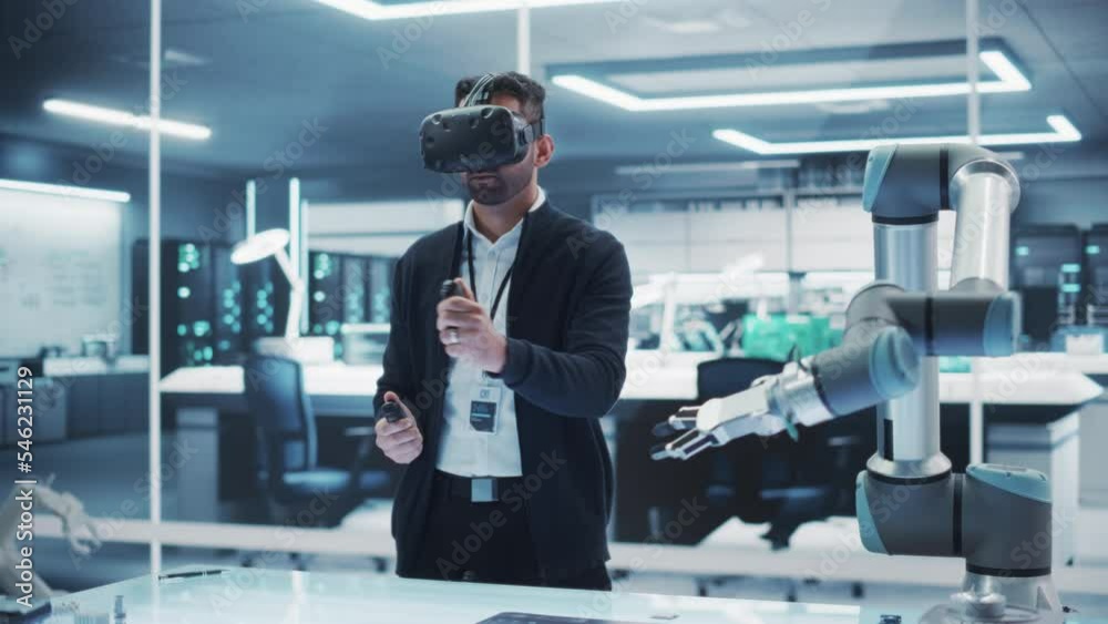 Portrait of a Young Specialists Using a Virtual Reality Headset with Controllers to Operate an Experimental Robotic Arm Machine. High Tech Industrial Laboratory Facility. Portrait of a Young Specialists Using a Virtual Reality Headset with Controllers to Operate an Experimental Robotic Arm Machine. High Tech Industrial Laboratory Facility.