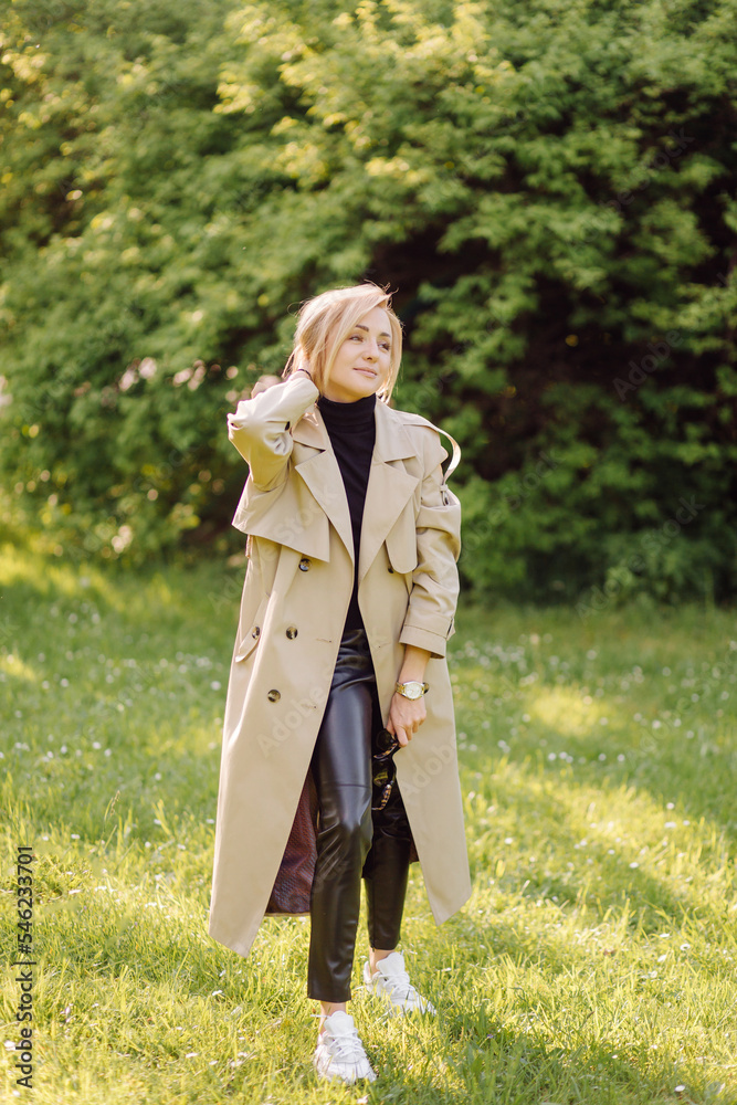 Caucasian blonde woman wearind trench smile happily on sunny spring day outside walking in park Caucasian blonde woman wearind trench smile happily on sunny spring day outside walking in park