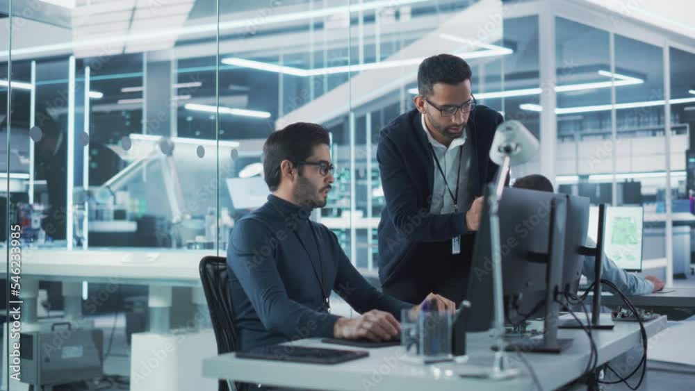 Two Industrial Engineers Discussing Work Projects, Talk and Use Desktop Computer in Robotics Startup Facility. Specialists and Professionals Researching and Developing High Tech Robotics Equipment. Two Industrial Engineers Discussing Work Projects, Talk and Use Desktop Computer in Robotics Startup Facility. Specialists and Professionals Researching and Developing High Tech Robotics Equipment.