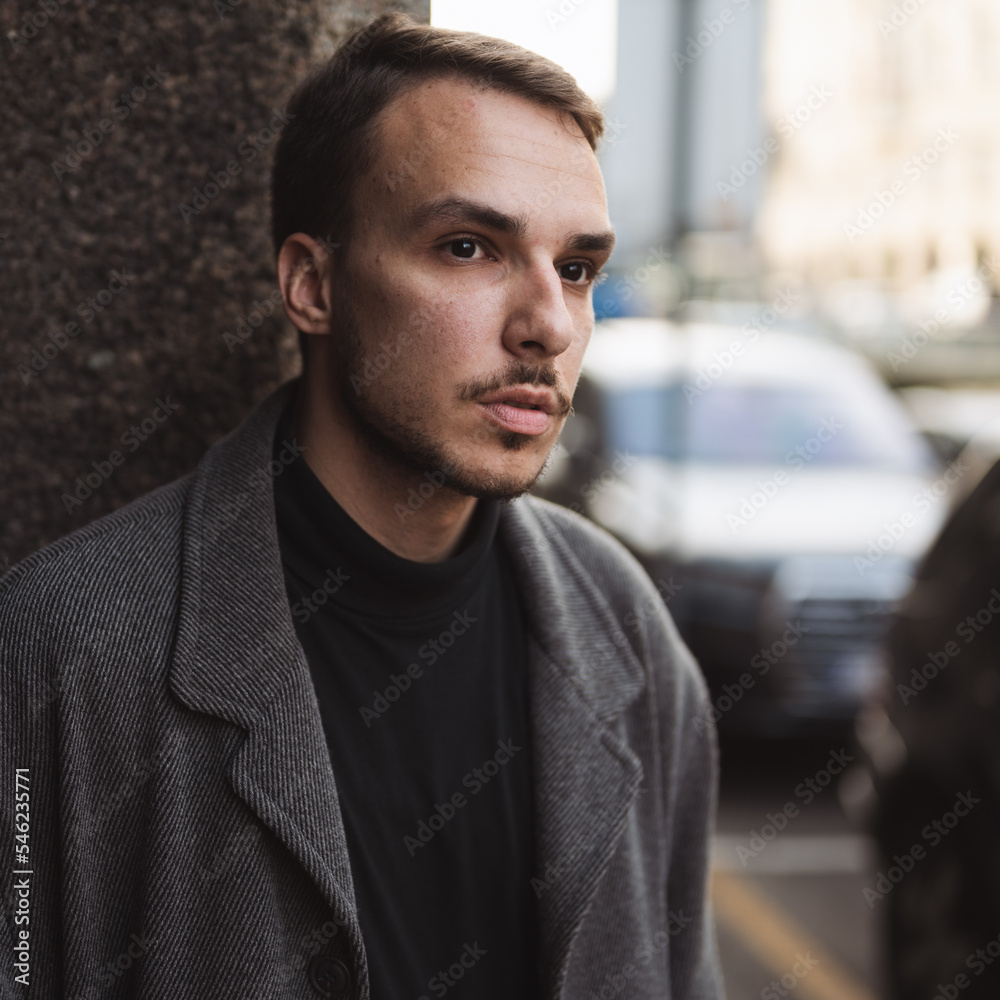 Handsome fashionable guy waiting for a taxi on street. Head and shoulders portrait of young man in dark coat looking at camera right with parked cars in background
