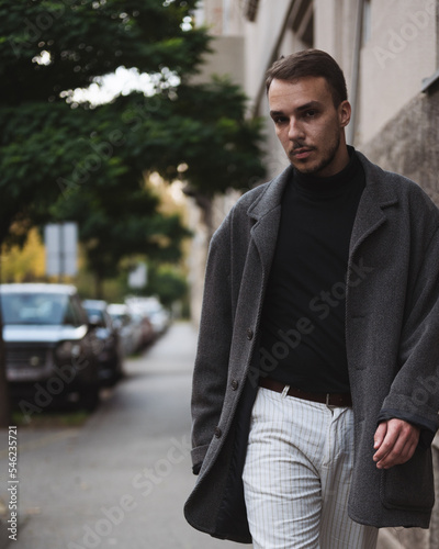 Fashionable attractive young man in dark coat and white trousers walking toward camera with self-confidence. Strong body language, city street fashion