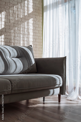 Sofa near wall in interior of living room. Closeup of soft comfortable gray sofa bed with cushion. Morning sunlight shining in showing beautiful window frame shadow. Background, Mockup, Chaise lounge