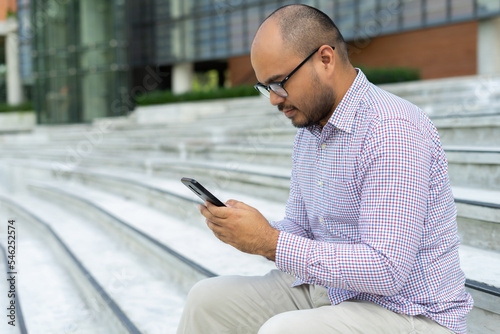 Bearded businessman smiling wearing glasses using modern smartphone device typing text message in social media while sitting on stairs outdoor. Happy male spending free time in break of working. © Patcharanan