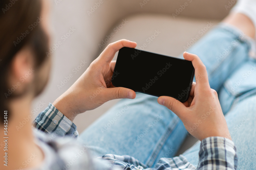 Young european male watches video, plays in game on smartphone with blank screen on sofa in living room Young european male watches video, plays in game on smartphone with blank screen on sofa in living room