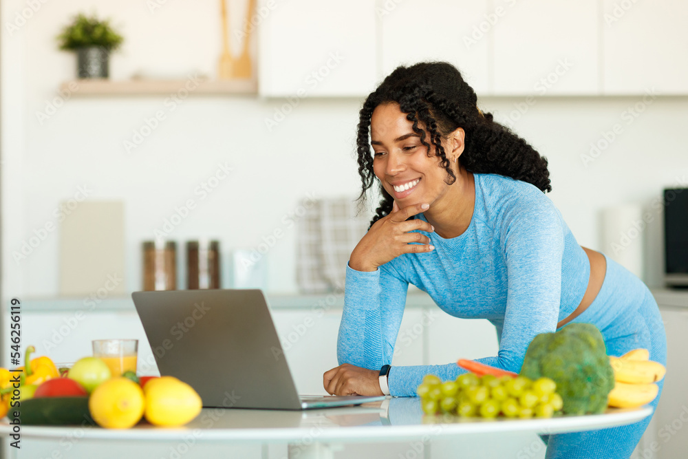 Excited black lady using laptop in kitchen, searching online healthy recipes while cooking lunch after domestic workout Excited black lady using laptop in kitchen, searching online healthy recipes while cooking lunch after domestic workout
