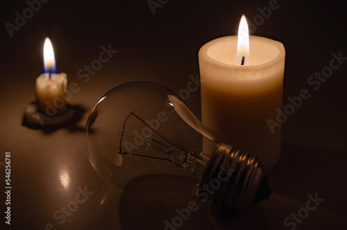 Switched off light or not glowing light bulb near a burning candles in total darkness. Blackout city, electricity off, load shedding, energy crisis or power outage, concept image. 